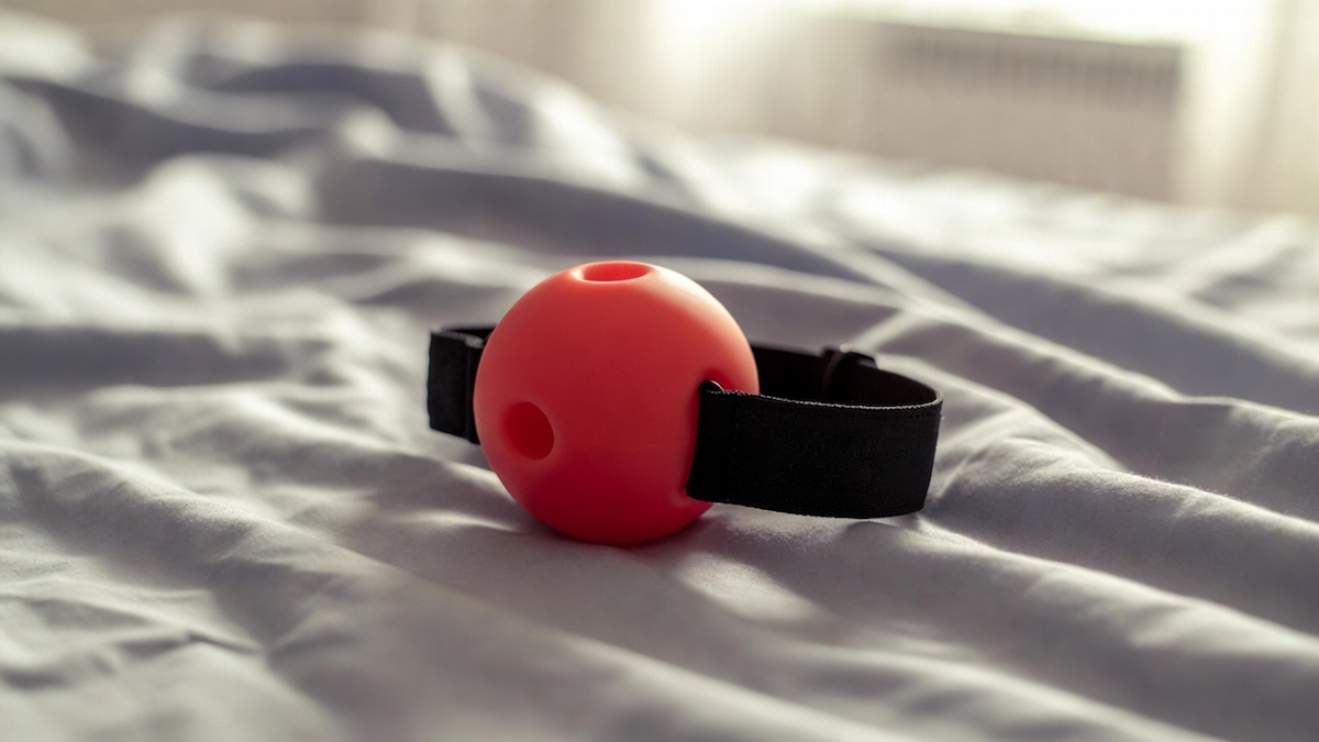 Ballgag con sfera rossa e cinturino nero appoggiata su un letto.